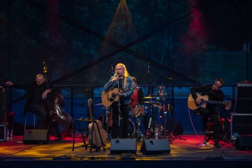 Andrzej Sikorowski na scenie podczas koncertu. Artysta stoi z gitarą. Po jego prawej znajduje się drugi gitarzysta a po lewej kontrabasista.