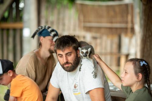 Mężycznza z lemurem na ramieniu, obok siedzi dziewczynka i głaszcze lemura