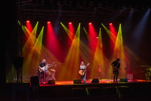 Wokalistka na scenie z gitarą. Po bokach gitarzyści. Światło żółto-czerwone.