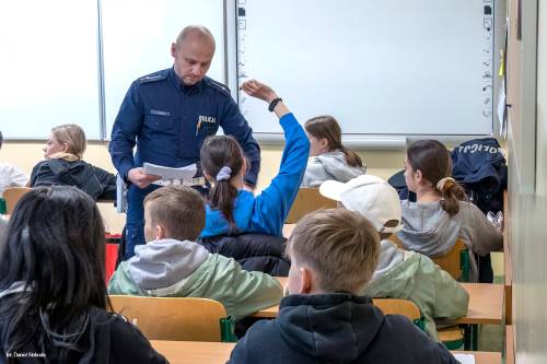 Rozpoczęcie Turnieju o bezpieczeństwie w Ruchu Drogowym - część teoretyczna. Organizatorzy stojący pod tablicą, uczestnicy siedzący w ławkach. rozdaje uczestnikom testy.