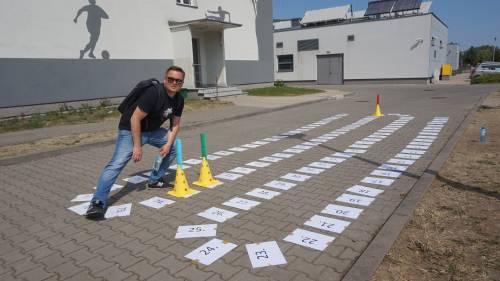 Nauczyciel pozuje na planszy do gry terenowej. Kartki z numerami ułożone na kostce brukowej.