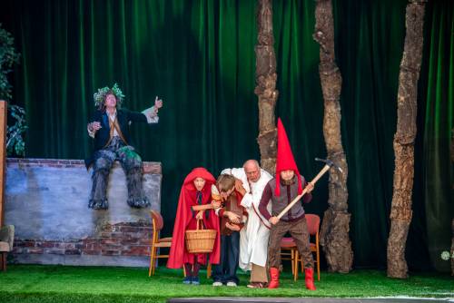 Czerwony kapturek, krasnoludek i dwie inne postacie na scenie. Aktorzy schylają się. Postać z nogami zwierzęcia siedzi na murze.