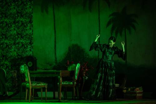 Aktorka w ciemnej, długiej sukni tańczy przy stole. Scena skąpana jest w zielonym świetle.