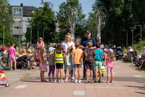 Grupa dzieci na deptaku słucha policjanta i kobiety, którzy stoją przed nimi.