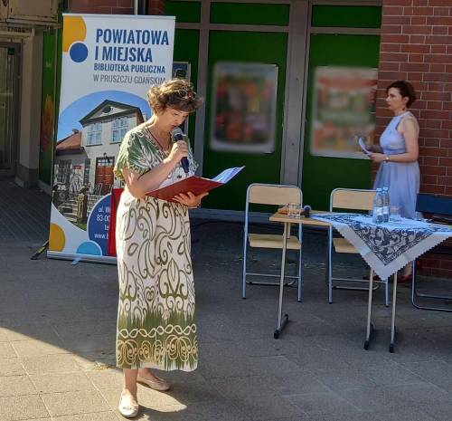 Kordiana czyta Zastępca Dyrektora Biblioteki. W tle inna uczestniczka i rollup biblioteki.