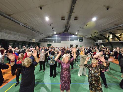 Seniorzy tańczą z uniesionymi rękami. Widać dużą grupę ludzi.