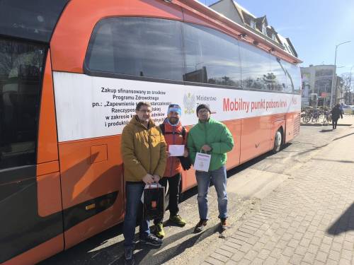 Trzech mężczyzna w kurtkach stoi przed czerwonym autobusem - mobilny punkt krwiodawstwa.