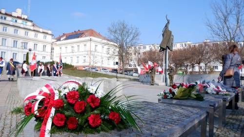 Uczestnicy obchodów zebrani na Placu Jana Pawła II w Pruszczu Gdańskim. Na pierwszym planie kwiaty leżące na ławkach.