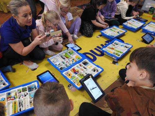 Grupa dzieci siedząca na macie z zestawami kloców i tabletami. W rogu widać instruktorkę.