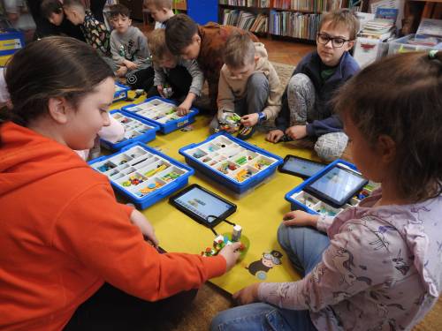 Grupa dzieci siedząca na macie z zestawami kloców i tabletami. Widok zza pleców.