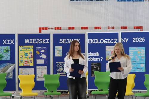 Dwie uczennice stoją przed tablicą z napisem XIX Miejski Sejmik Ekologiczny