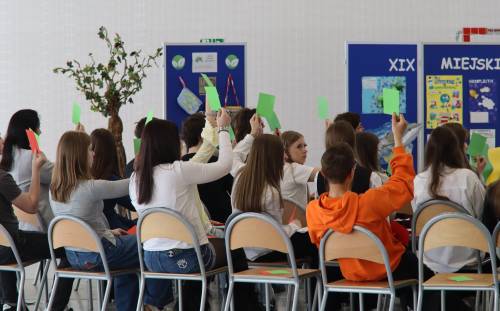 Grupa uczniów siedzi na krzesłach i trzyma podniesione kartki w dłoniach