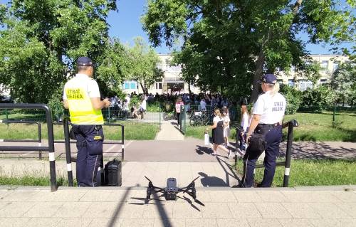 Strażnik i strażniczka miejska stoją z dronem przed wejściem do szkoły