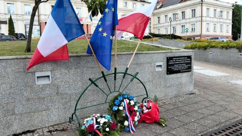 Zbliżenie na wiązanki kwiatów, flagi oraz tablicę pamiątkową