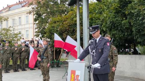 Lotnik w mundurze stoi przy przed pomnikiem, w tle żołnierze