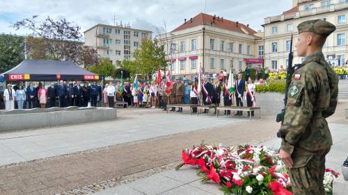Widok na żołnierza i wiązanki kwiatów oraz ludzi zgromadzonych na placu