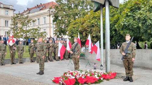 Trzech żołnierzy stoi pod pomnikiem, w tle grupa żołnierzy