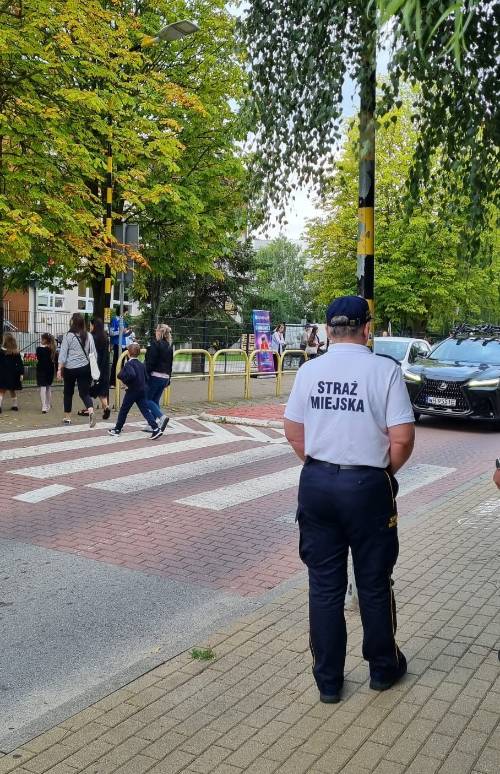 Strażnik miejski stoi na chodniku przy przejściu dla pieszych, w pobliżu którego przechodzą dzieci.