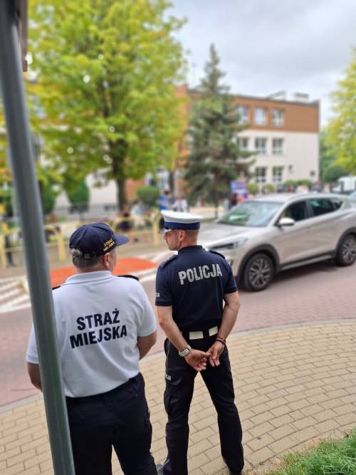 Strażnik miejski oraz policjant stoi na chodniku przy przejściu dla pieszych, w pobliżu którego przechodzą dzieci.