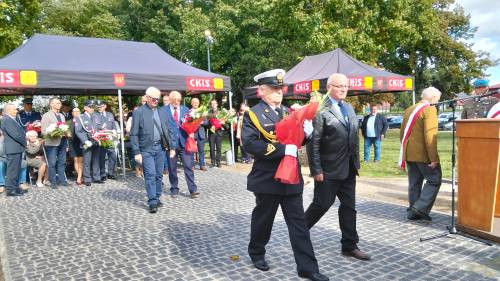 Zbliżenie na dwóch panów idących złożyć kwiaty, z tyłu inni uczestnicy uroczystości
