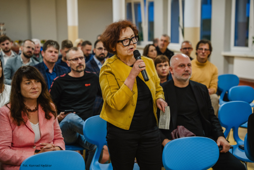 Kobieta z mikrofonem w ręku, obok siedzą inni uczestnicy spotkania